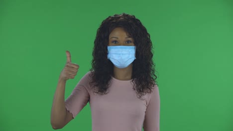 portrait of beautiful african american young woman in medical protective face mask looking at camera and showing thumbs up gesture with both hands. burning brunette with wavy hair in jeans and a beige blouse on a green screen in the studio. health