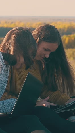 pretty girl with laptop looks into friend tablet in camp on steep river bank in early autumn morning slow motion