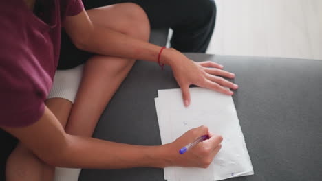 child seated on sofa with leg crossed writing on paper using pen with visible partial presence of person in black trouser beside her in relaxed indoor setting showing creativity and learning moment