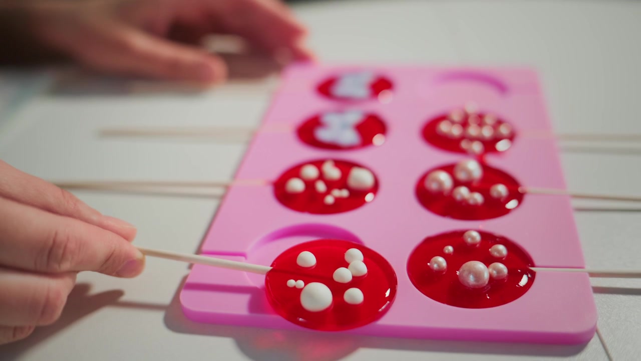 Hands carefully insert sticks into red lollipop molds. Red candy gels with white pearl sprinkles. Close-up of sweet-making process. Ideal for food, dessert, or crafting videos.