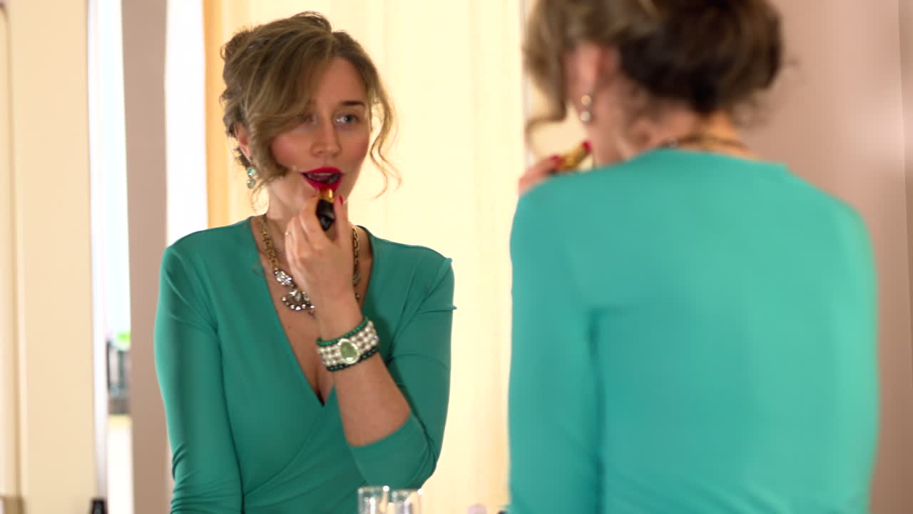 Woman in turquoise dress putting on red lipstick in front of a mirror