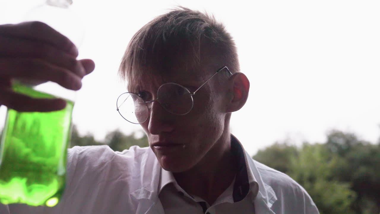 Portrait view of botany student in white coat sitting outdoors chewing snack and sipping lemonade drink while looking aside with relaxed expression during break near greenery and wet pavement