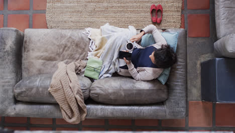 Overhead-view-of-biracial-woman-lying-on-sofa-under-blanket-using-smartphone-at-home,-slow-motion