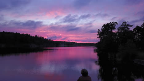 aerial backward drone shot, bypassing a man sitting at a rocky shore, of a lake, a purple sky, a colorful sunset or dusk, at albysjon, tyreso, sweden