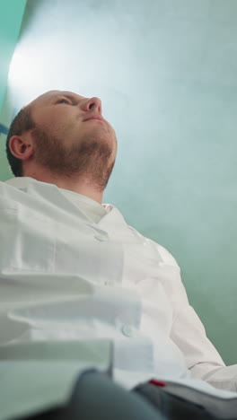 a downward view of a technician in a white lab coat, sitting and looking up thoughtfully in a well-lit laboratory with light shining from the desk lamp and microscope and on the wall