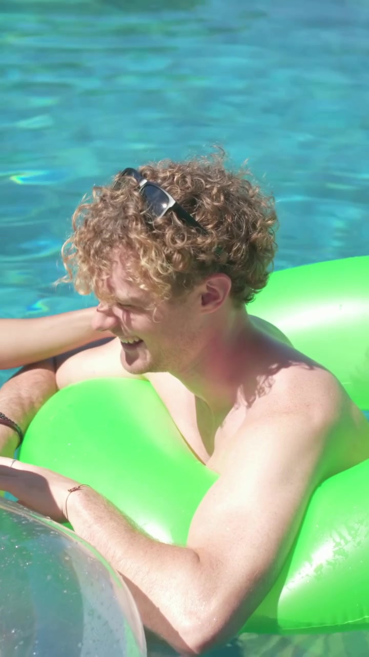 Vertical video: Smiling shirtless man in shades responding to reaching hand, on green float in pool