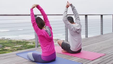 Happy-diverse-couple-doing-yoga,-sitting-and-stretching-on-balcony