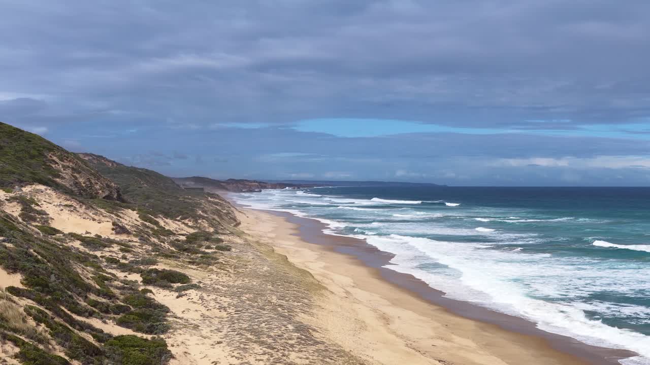 Drone glides above sandy coastline, capturing waves, dunes, and coastal vegetation in daylight