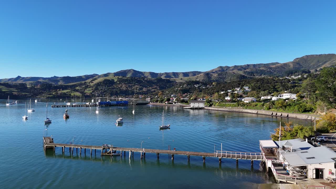 Aerial footage captures Akaroa's lighthouse and harbor under clear skies, showcasing serene waters and anchored boats