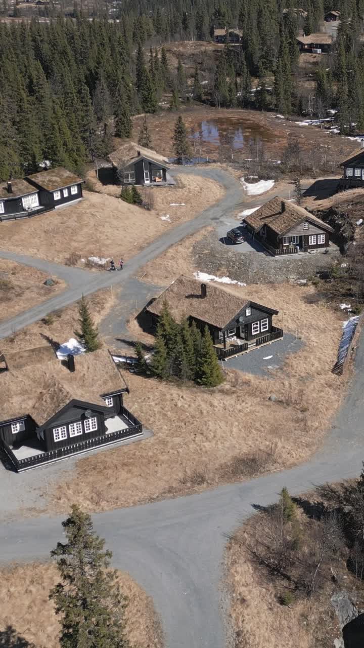 Drone vertical shot showcases Danebu Kongsgaard, a mountain lodge in Valdres, Norway. Wooden houses with grass-covered roofs, surrounded by forests and scattered snow under a bright, sunny sky.