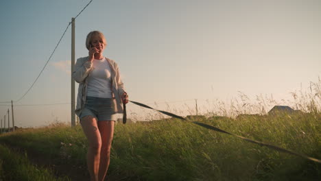 dueño de perro en pantalones cortos de jean en llamada telefónica caminando a lo largo de un campo de hierba al amanecer, sosteniendo la correa mientras el perro tira hacia adelante enérgicamente, poste eléctrico y edificio lejano visible en el fondo