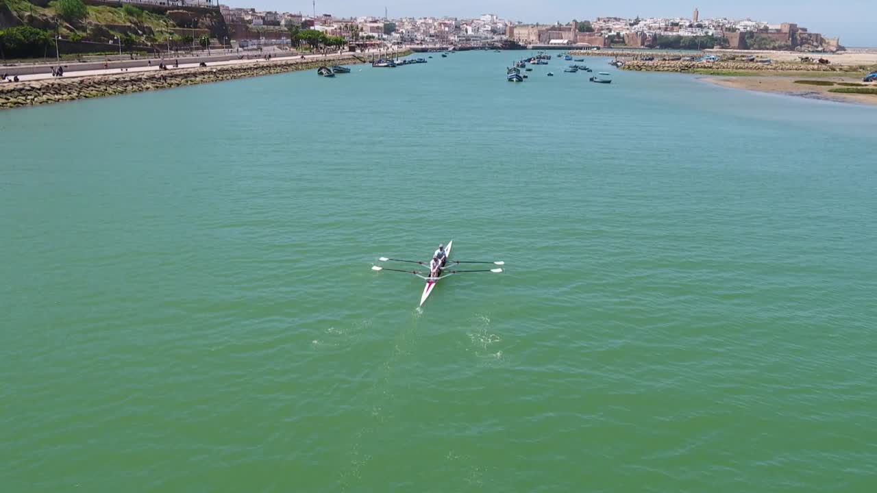 Cinematic drone shot following two rowers in a boat on Bou Regreg river. Features synchronized paddling action with the historic Kasbah of the Udayas walls and blue sky in the background