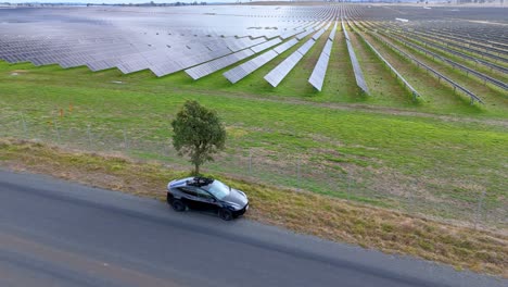a modern electric vehicle travels along a rural road beside an expansive solar panel array under daylight, captured in a smooth aerial tracking shot