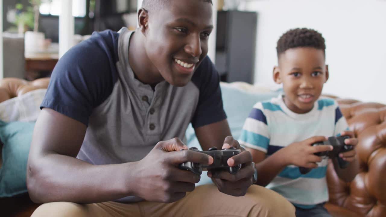 Video of happy african american father and son sitting on sofa and playing video games