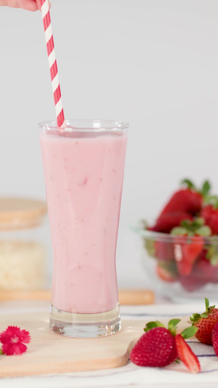Una mano inserta una pajita a rayas en un cremoso batido de fresa, con iluminación de estudio brillante y un fondo de frutas frescas.