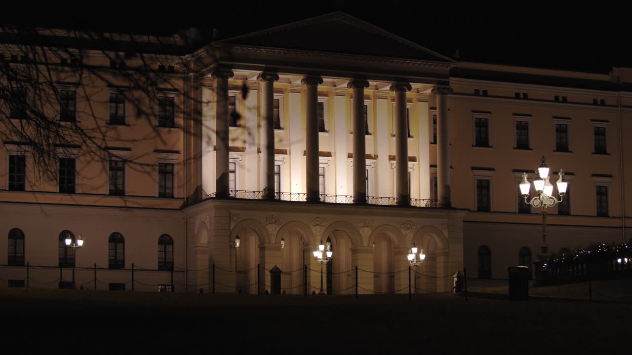 Slow motion wide 4K shot with parallax motion of branches from tree in front of illuminated front entrance pillars of the Norwegian Royal Palace on top of Karl Johan street, at night in Oslo Norway