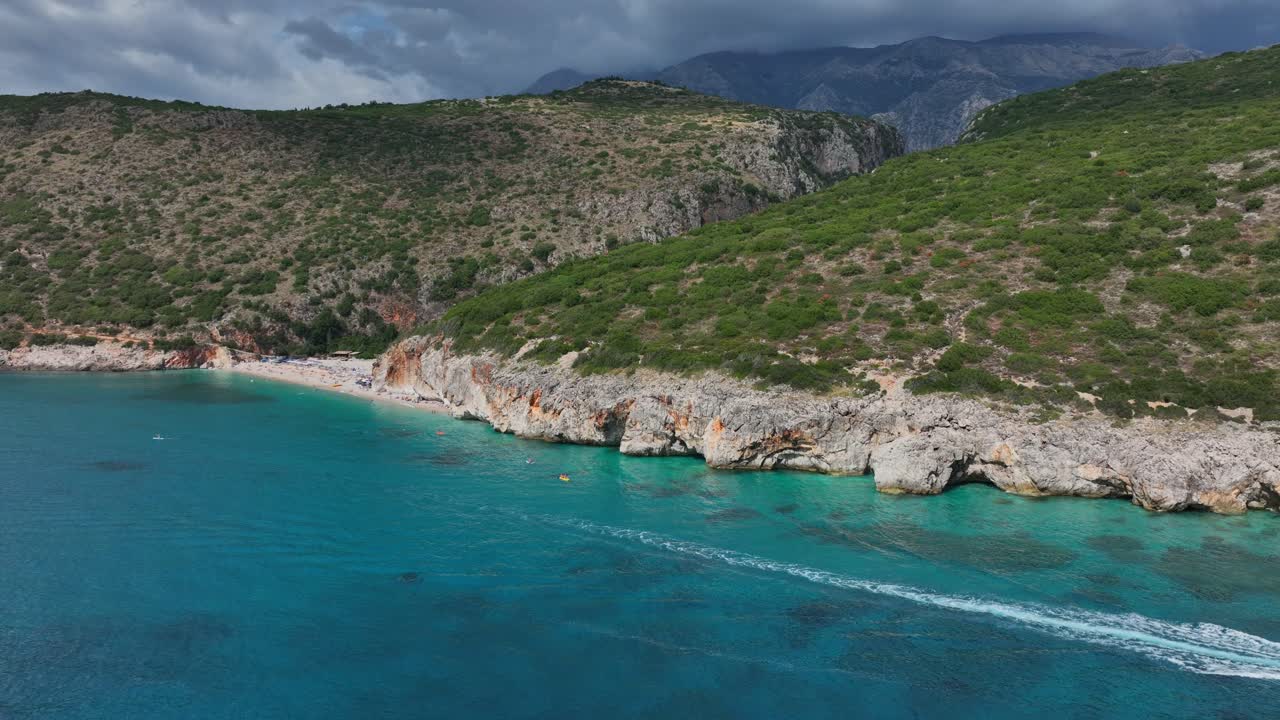 Sunny day aerial of boat crossing Gjipe Beach showing waves lapping rocky coast and surrounding cliffs