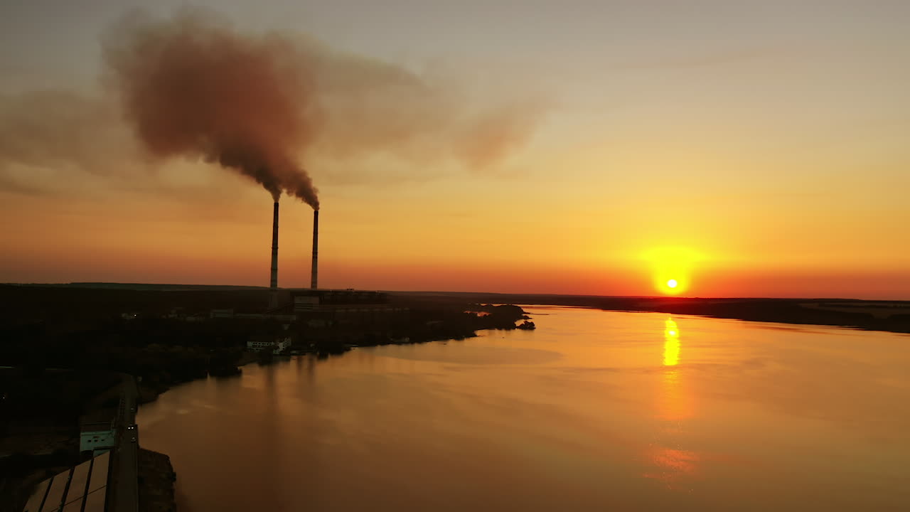 Industry near the river in the evening. Factory pipes release smoke into nature at sunset time. Pollution the environment. Aerial view. Camera moves back.