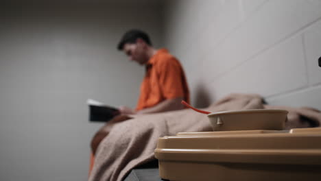 A-convict-in-prison-cell,-prisoner,-inmate-in-orange-prison-jumpsuit,-uniform-reading-a-book,-bible-in-his-jail-cell