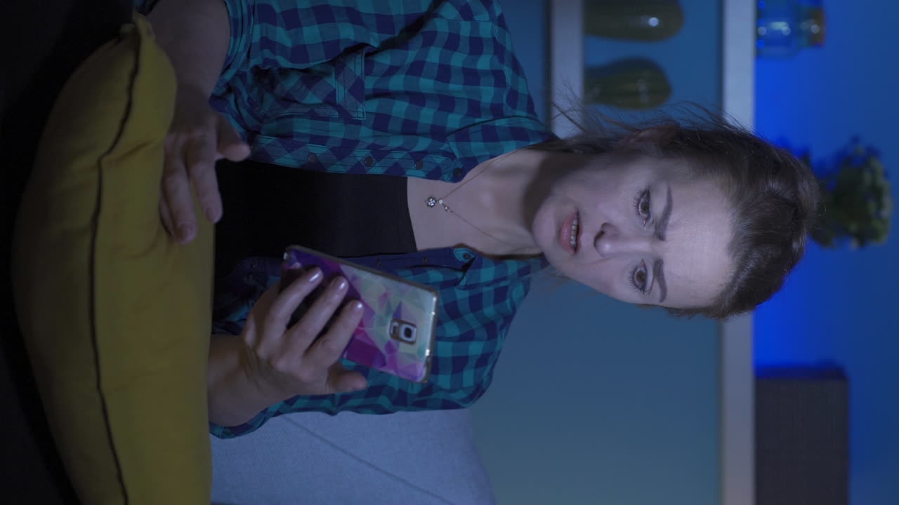 밤에 집에서 전화로 문자 메시지를 보내는 여성의 수직 비디오는 실망합니다.