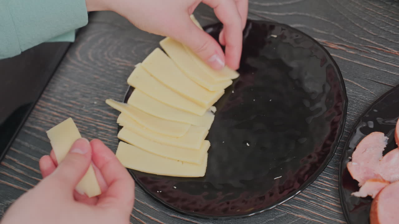 close up of chef placing thin yellow cheese slices in neat row on black ceramic plate over wooden table surface in kitchen, with partial view of sausage slices on side plate and hand visible in frame