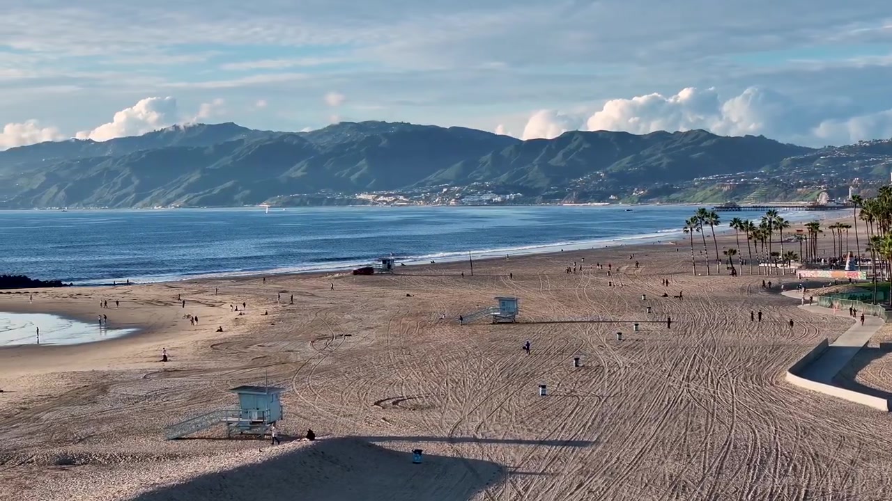 Toma aérea cinematográfica de dron de 70mm girando lentamente hacia adelante sobre Venice Beach y la costa del Pacífico de Los Ángeles, con las montañas de Santa Mónica elevándose en el fondo distante.