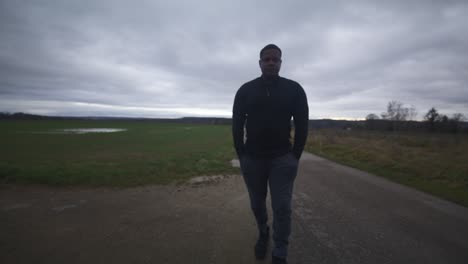 modern black male waiting in open field under grey sky, europe, 4k | muscular, intimidating, shadow, figure, blurry, dark clothes, powerful, walk towards, serious, looking