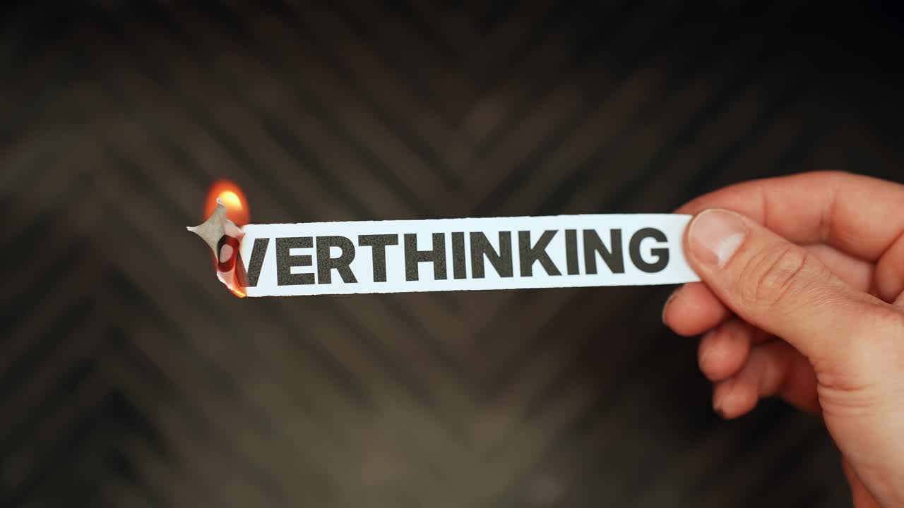 Hand holds a paper strip reading “OVERTHINKING” while the edge burns in flame, symbolizing anxiety, mental overload, and intrusive thoughts