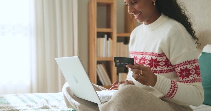 African American woman sitting on bed in white sweater typing on laptop, checking card to pay