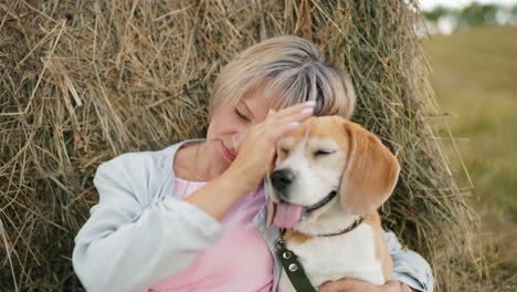 woman holds her dog closely, gently touching its ear affectionately while resting against hay bale in vast farmland, she looks at her dog and rests her head on its head