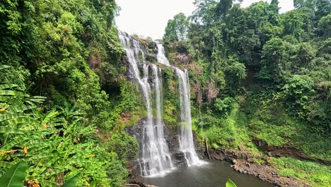 a wide shot captures a lush tropical jungle waterfall in laos, with a zipline crossing the scene. bright daylight, vibrant greenery, and steady camera movement enhance the adventurous mood