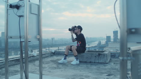 Offene-Aufnahme-Eines-Jungen-Asiatischen-Fotografen,-Der-Am-Frühen-Morgen-Auf-Dem-Dach-Sitzt-Und-Die-Innenstadt-Der-Stadt-Einfängt