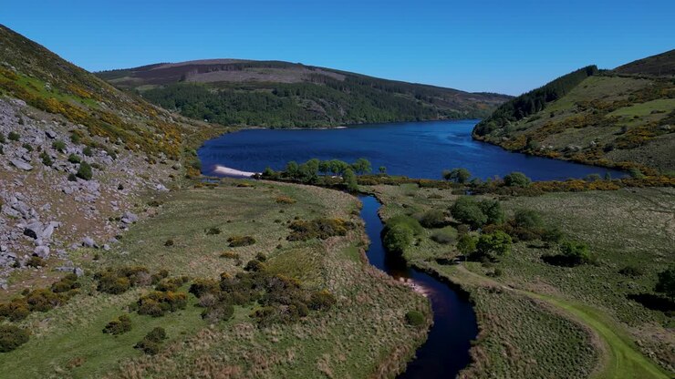 Vista aerea in movimento di un drone che avanza su un fiume tortuoso che sfocia nel Lough Dan, circondato da colline e aperta campagna a Wicklow, Eire, catturando la bellezza naturale in una luminosa giornata di sole