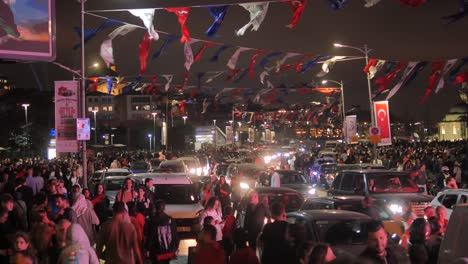 a large crowd of people gathers on a busy street at night in a city. the street is decorated with flags and lights. there are many cars in the traffic.