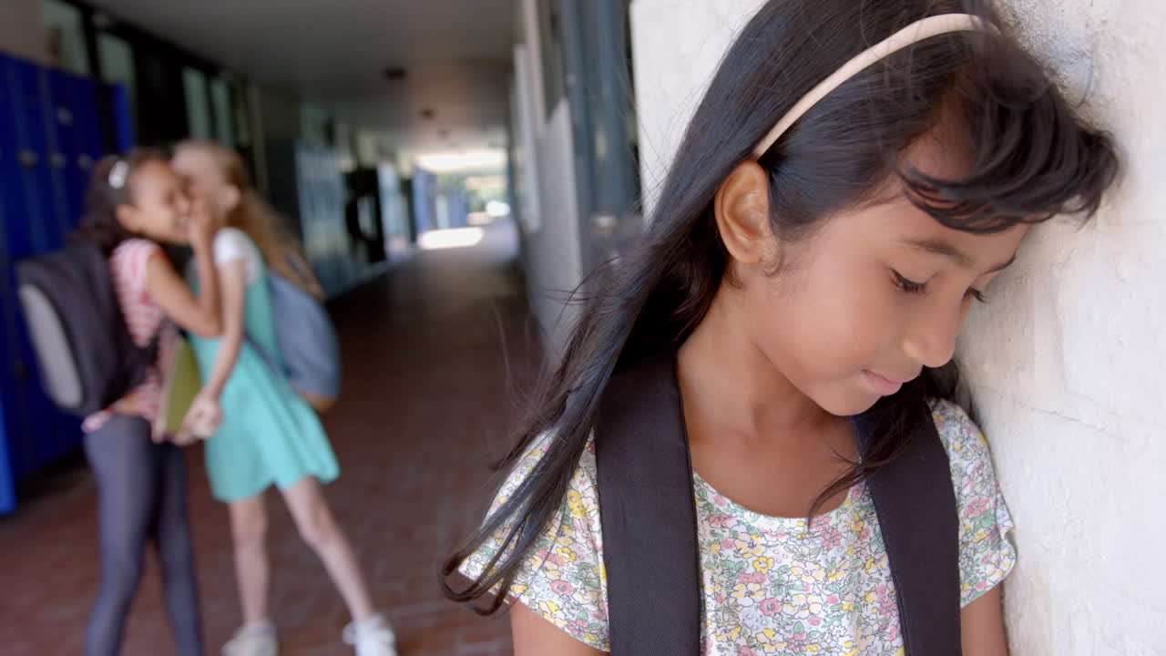 In school, girl with backpack feeling sad while other girls whispering nearby