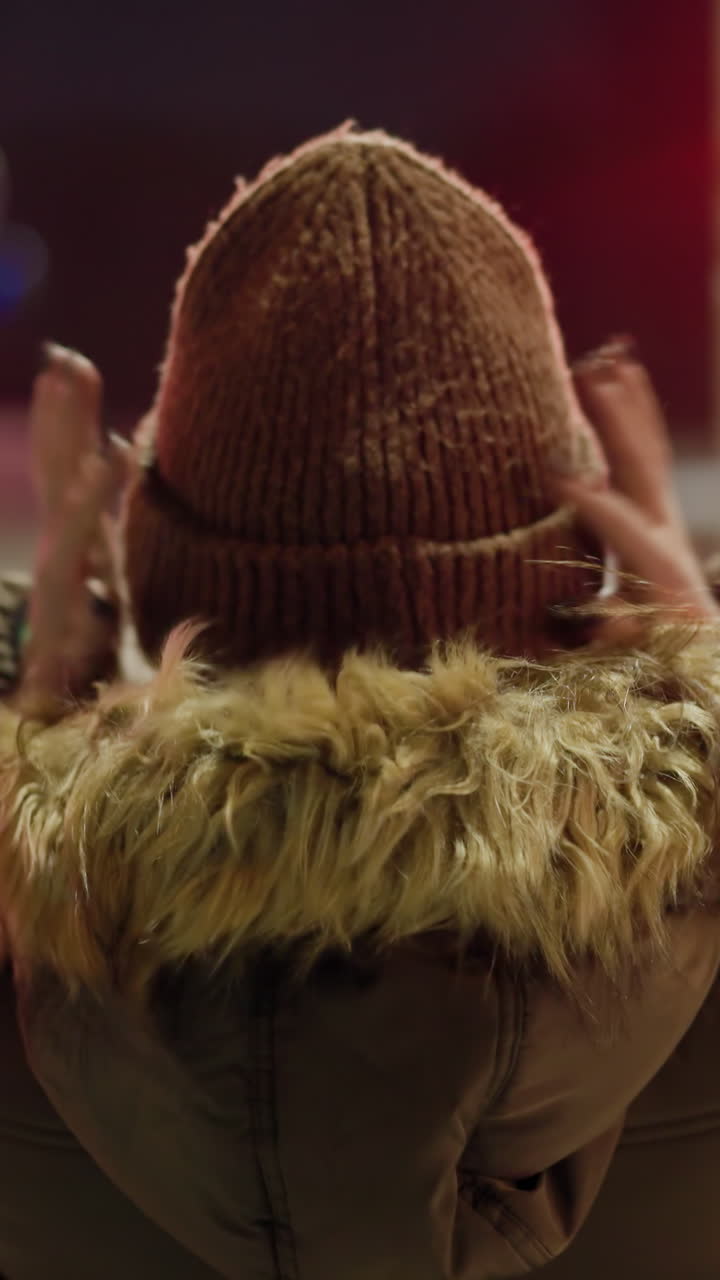 First-person view of a woman in a winter coat with a fur hood, holding her head with both hands, standing at a nighttime urban intersection with blurry lights and red traffic signal in the background