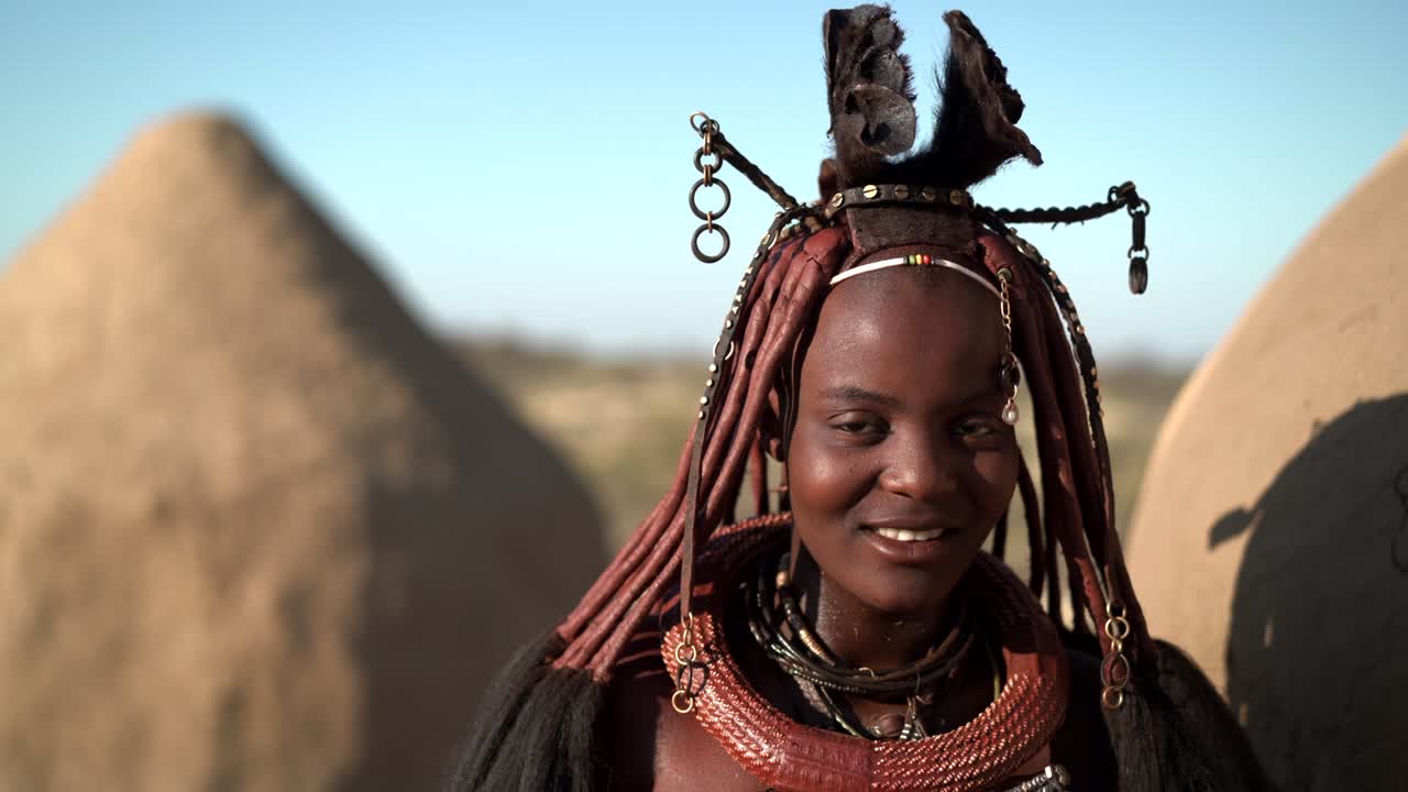 una joven himba feliz sonriendo, de pie junto a una cabaña en el tradicional pueblo himba cerca de kamanjab en namibia, áfrica