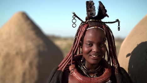 happy young himba woman smiling, standing next to hut in traditional himba village near kamanjab in namibia, africa