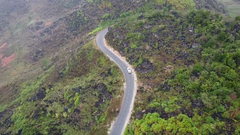 Drone video captures a solitary car on a winding mountain road in Vietnam. The lush greenery and rugged terrain create a serene, untouched atmosphere under soft sunlight, with no people visible