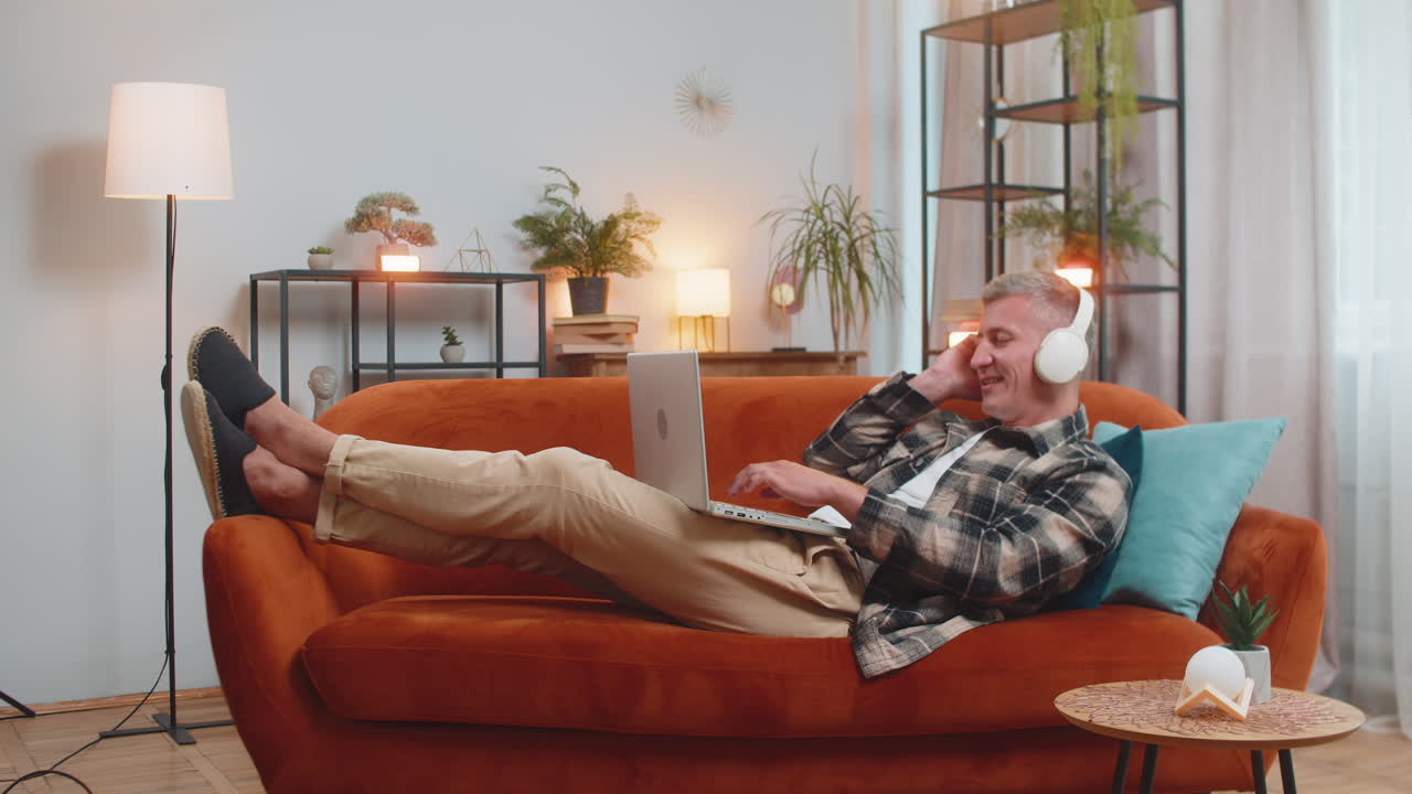 무선 헤드폰을 착용한 행복한 남자가 집 소파에 누워서 음악을 듣고 노트북에서 일하며 휴식을 취하고 있습니다.