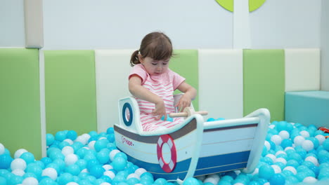 excited girl toddler sitting in toy boat and tries to catch balls from dry pool which mom throw in kids playroom