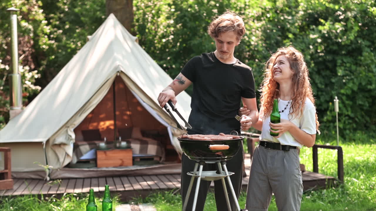 Young couple frying meat on the grill and drinking beer, laughing. Greenery around. Glamping. Slow motion