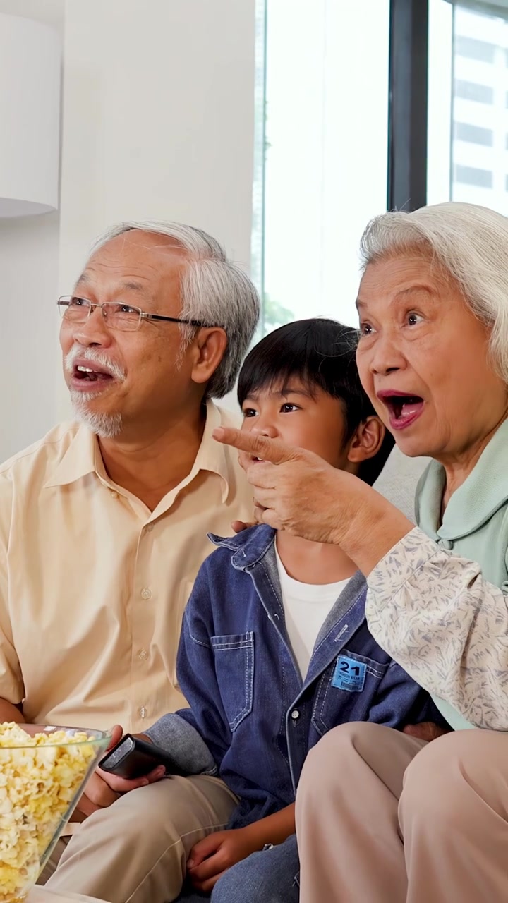 明るいリビングルームで、三世代のアジア系家族がポップコーンを分け合い、笑いながら一緒に映画を観ている。