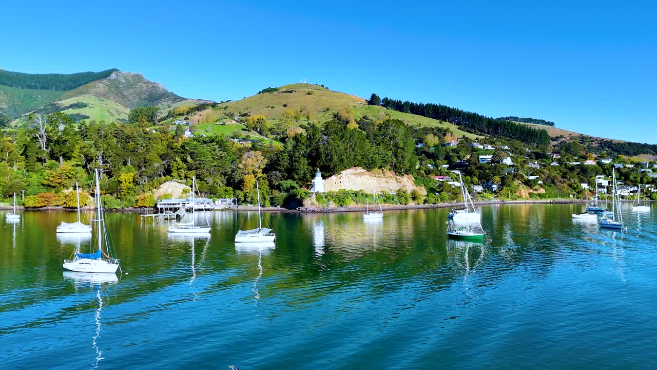 Aerial view of Akaroa Harbour with sailboats, lush greenery, and a lighthouse under clear blue skies