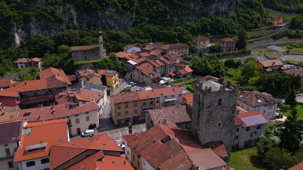 Aerial view of medieval stone tower and terracotta rooftops surrounded by green cliffs and churches. Drone forward descend. Shot in Cividate Camuno, Italy (Cividate Camuno, Italia)