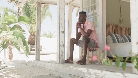 thoughtful african american man sitting on porch of wooden beach house looking away, slow motion