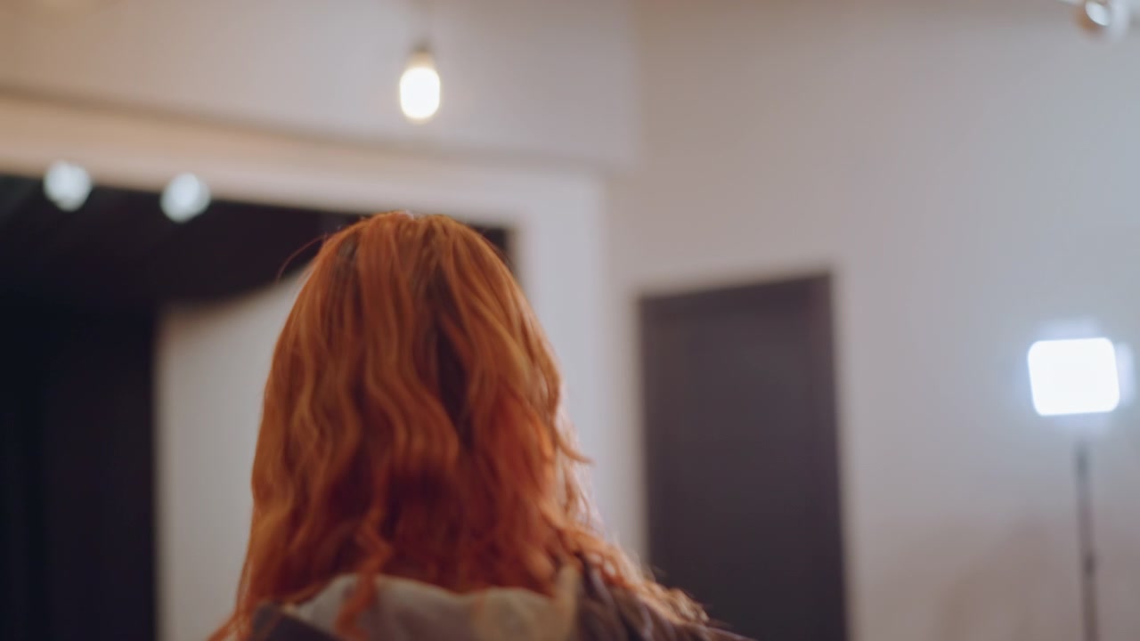 Cantante dai capelli rossi di fronte alle luci dello studio, vocalista nervosa che prova sotto una luce chiave brillante, studente con felpa con cappuccio riceve coaching vocale fuori campo, bagliore caldo della lampada, atmosfera tranquilla di una scuola di musica, primo piano.