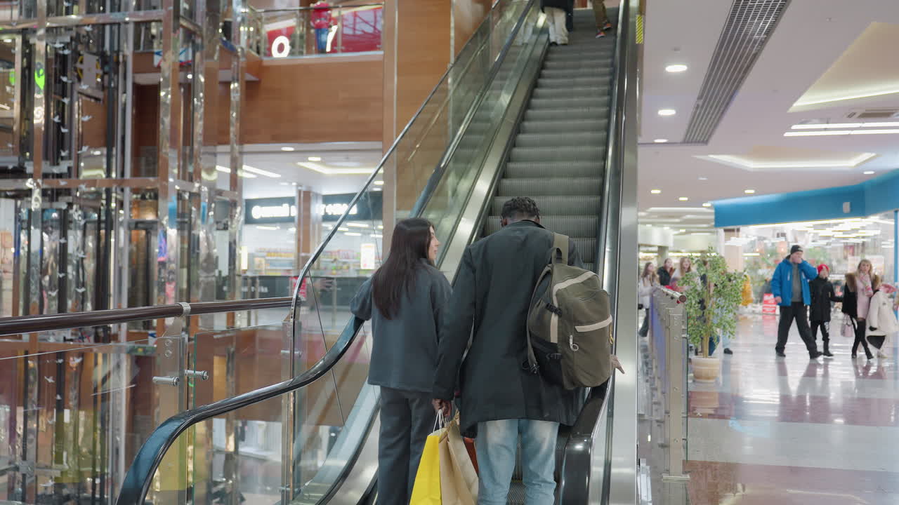 Rear view of young friends holding shopping bags while riding ascending escalator in modern indoor shopping mall, surrounded by glass railings, lights, and commercial space