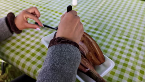 Close-up-of-hands-using-utensils-to-eat-a-grilled-fish-served-on-a-white-plate-at-a-green-checkered-picnic-table