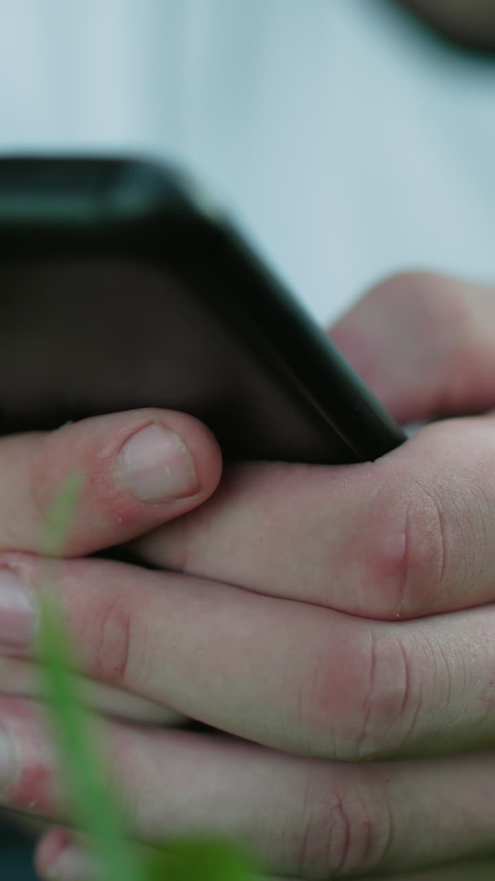 primer plano de un hombre con una camiseta blanca acostado en un campo cubierto de hierba mientras sostiene y opera un teléfono inteligente, centrarse en las manos agarrando el teléfono, hierba verde vibrante en un fondo borroso